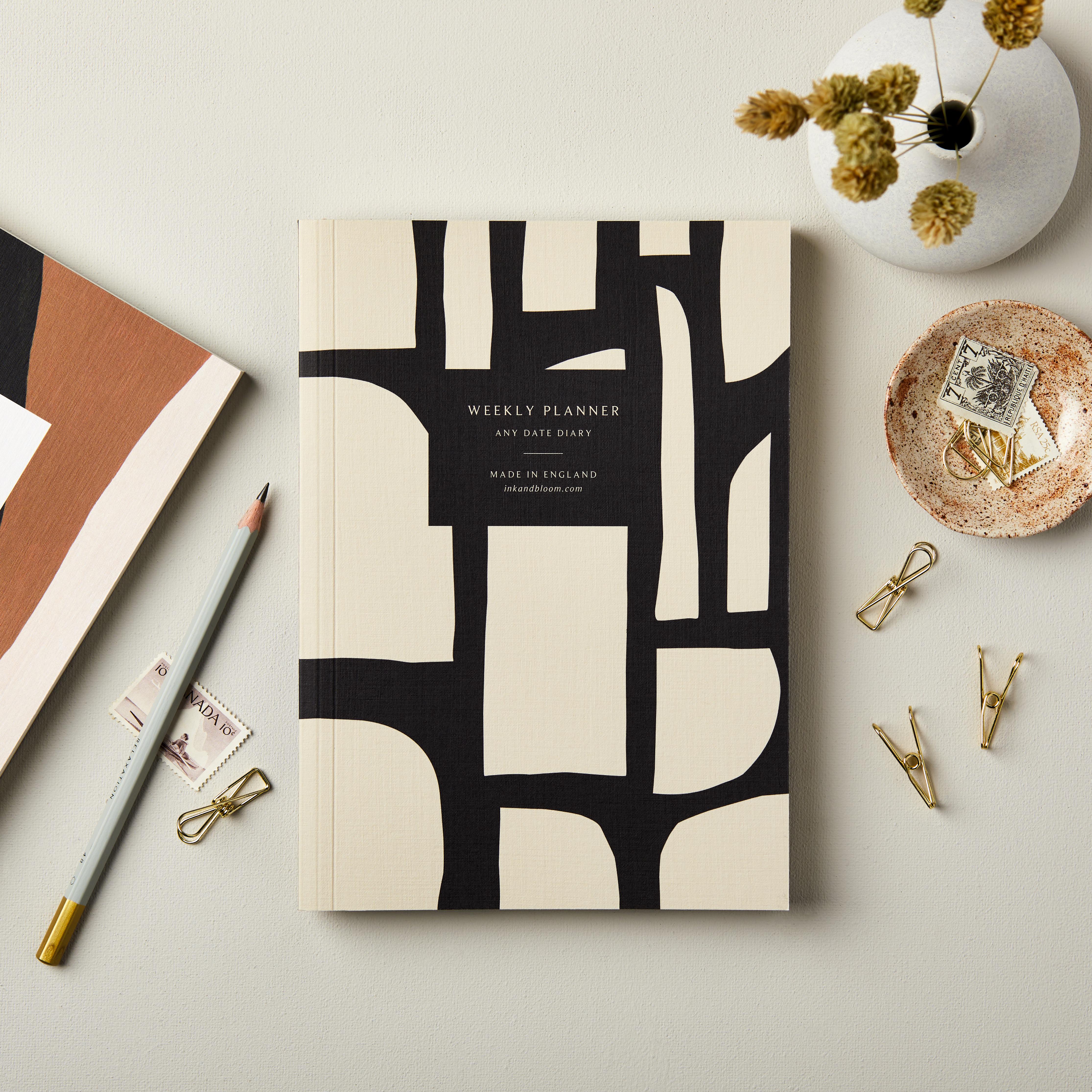 Weekly Planner,  A5, Undated with 'Any Date Diary' text, abstract black/cream design, on cream backdrop with vase and stationery.